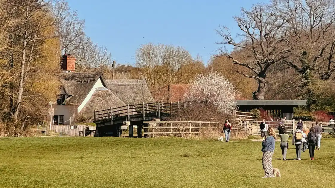 Walkers crossing Flatford Bridge near the thatched cottage in Dedham Vale National Landscape, with spring trees and open countryside under clear blue skies.