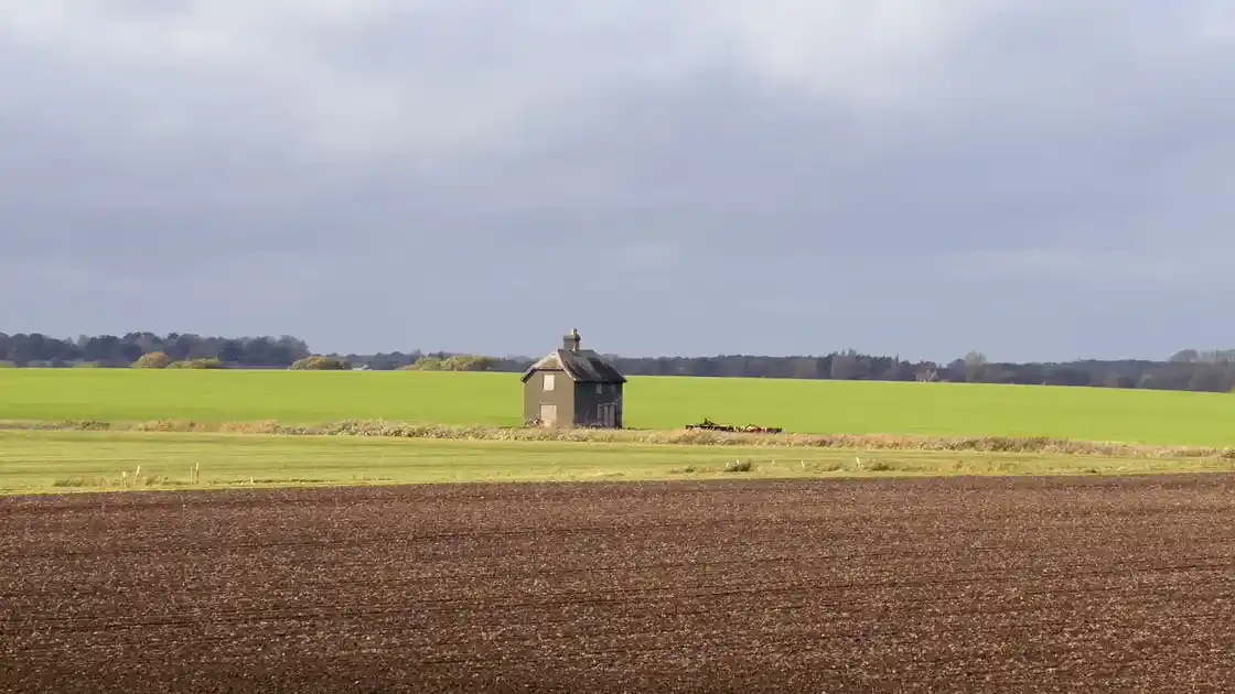 A lone, weathered house stands in open fields near the England Coast Path at Felixstowe Ferry, surrounded by wide farmland under a cloudy sky.