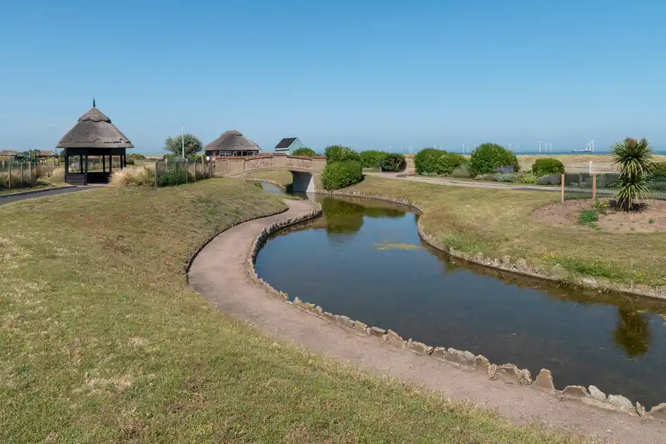 Walking path beside a coastal waterway with grassland and shelters near the Norfolk coast.