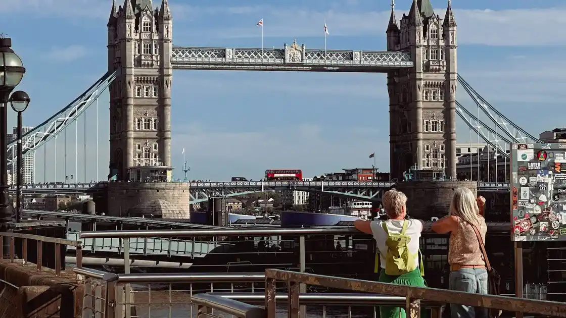 Two people standing by the River Thames looking toward Tower Bridge with a red London bus crossing.