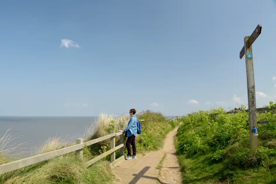 Walker on a coastal path beside the sea with a wooden waymarker on the East of England coast.