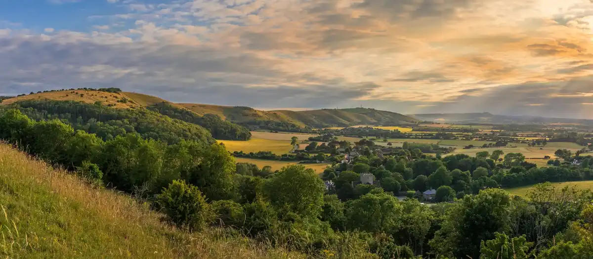 Panoramic view of the South Downs in South East England at sunset, with rolling chalk hills, farmland, and wooded valleys overlooking a rural village.