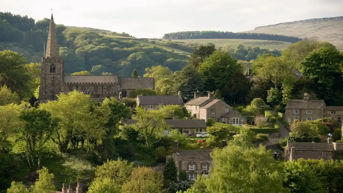 Aerial view of Hathersage village with St Michael and All Angels' Church spire rising amidst stone cottages and autumnal trees, set against the rolling hills of the Peak District in Derbyshire, England.
