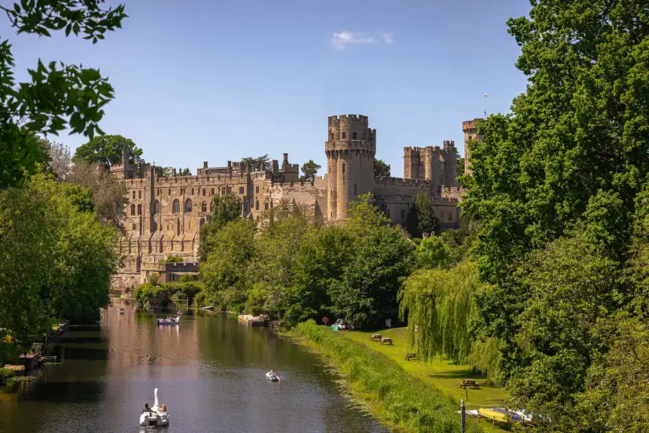 Warwick Castle overlooking the River Avon, with boats on the water and mature trees along the riverbanks in Warwickshire.