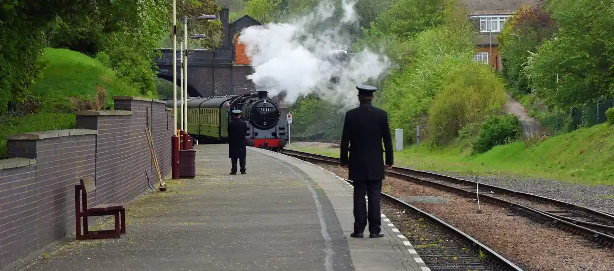 Heritage steam locomotive arriving at a railway station in Leicestershire, with railway staff on the platform and woodland surrounding the tracks.