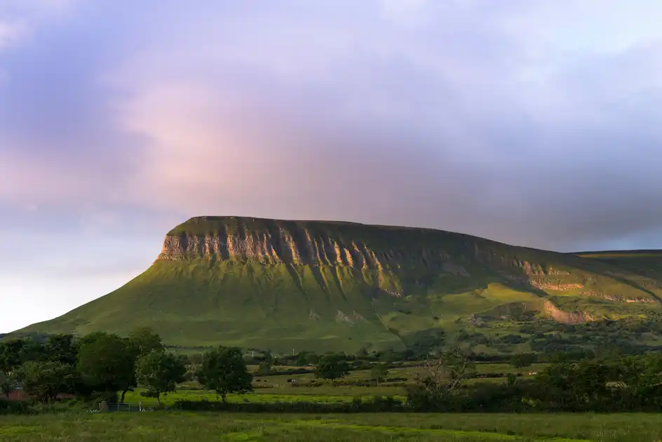 Benbulben mountain rising above green fields and woodland in County Sligo, Ireland, with evening sunlight illuminating its distinctive limestone cliffs.