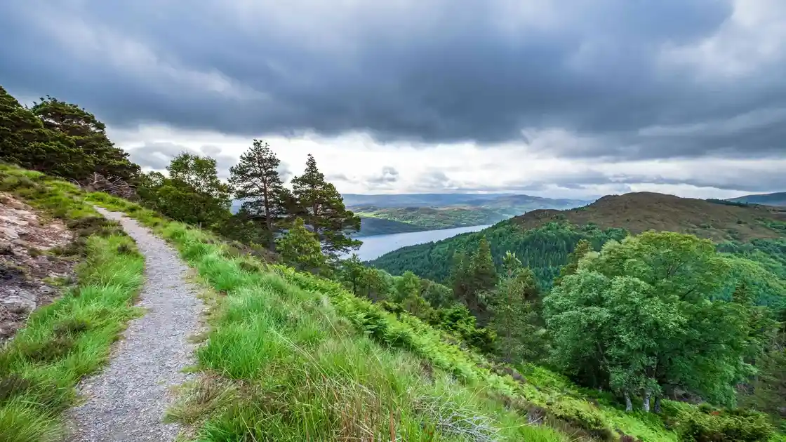 Hiking trail through the Scottish Highlands with views of a loch, forested hills, and dramatic skies above the glen.