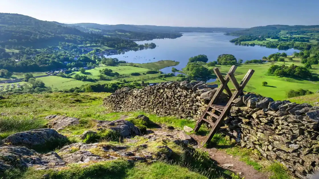 Traditional wooden stile crossing a dry-stone wall on a hillside walking path, overlooking a lake and green countryside in the Lake District, Cumbria.