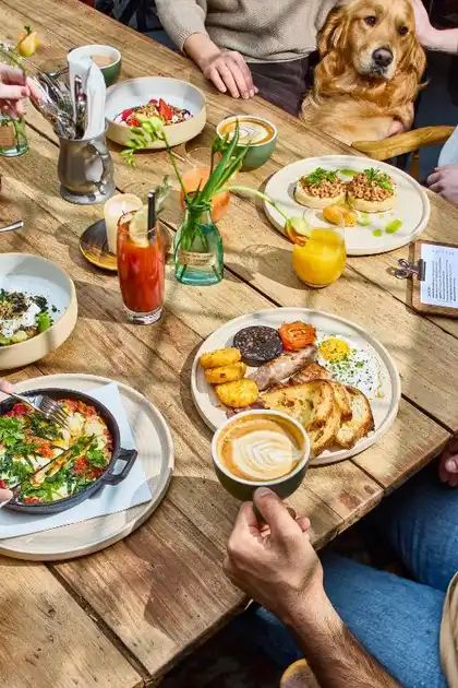Breakfast dishes, coffee and fresh juices served at a wooden table at The Chequers Inn in Thornham, Norfolk, with a dog seated alongside guests.