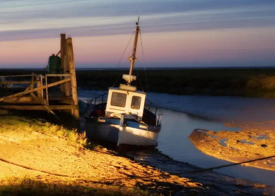 Fishing boat moored beside a wooden jetty at Thornham Harbour on the Norfolk coast, with tidal mudflats and a pastel evening sky.
