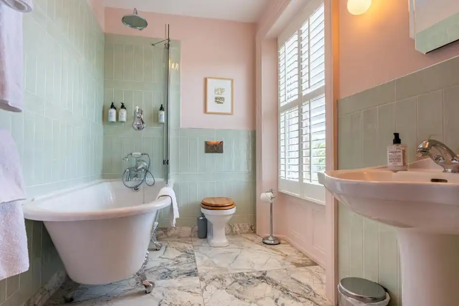 En-suite bathroom at Titchwell Manor Hotel in Titchwell, Norfolk, featuring a freestanding roll-top bathtub, overhead shower, pale green tiles and marble flooring.