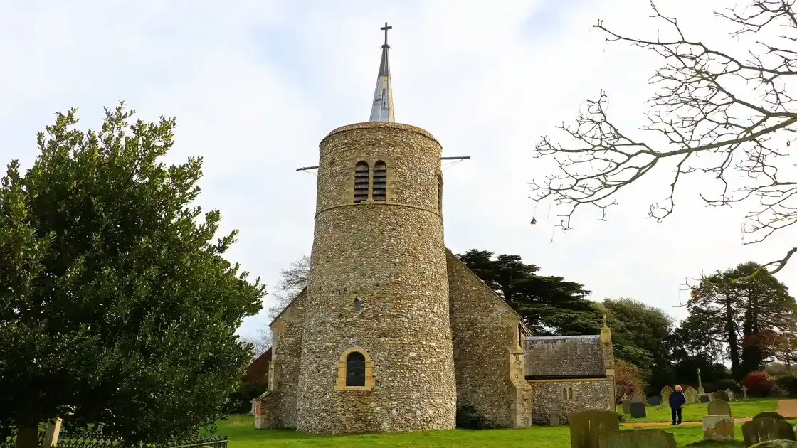 St Mary's Church in Titchwell, Norfolk, featuring a historic flint round tower, slender spire and churchyard with gravestones.