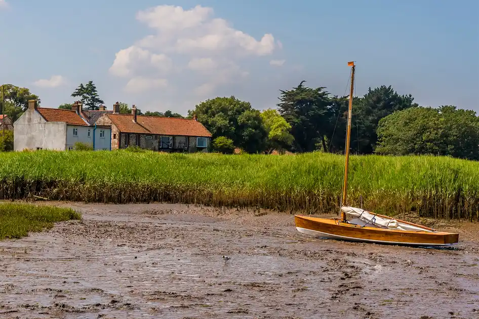 Small sailing boat resting on tidal mudflats near Brancaster beside a saltmarsh creek on the Norfolk coast, with traditional cottages and trees in the background.