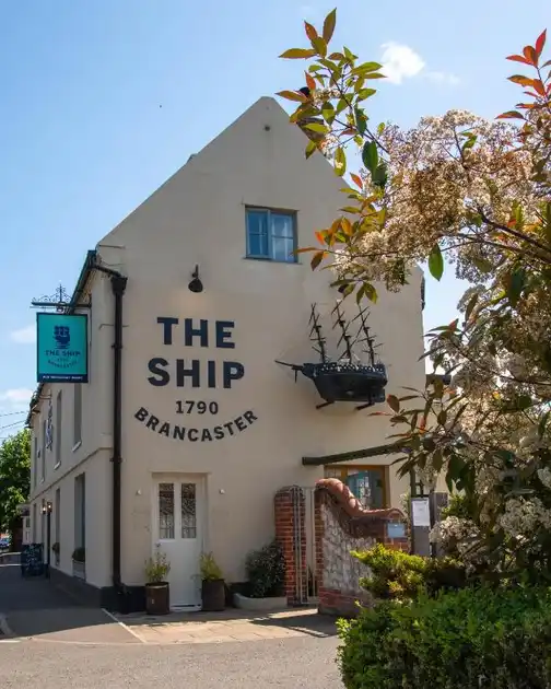 Exterior of The Ship in Brancaster, Norfolk, showing the whitewashed historic inn with its painted signage, entrance and hanging pub sign.