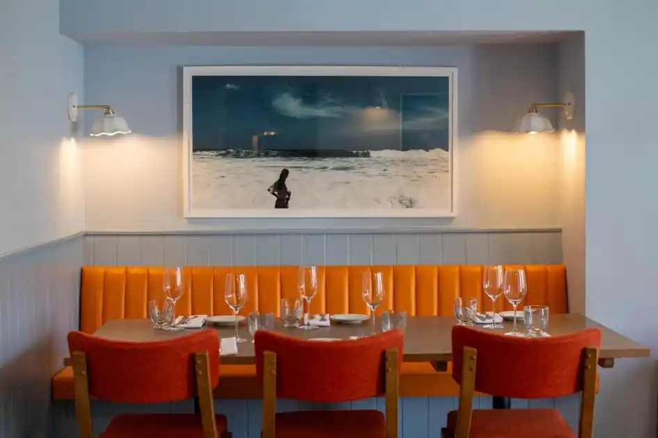 Restaurant dining area at The Ship in Brancaster, Norfolk, featuring orange upholstered seating, a wooden dining table, wine glasses and coastal artwork.