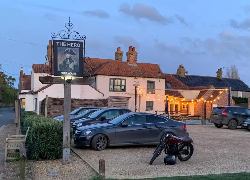 Exterior of The Hero in Burnham Overy Staithe, Norfolk, showing the traditional whitewashed village pub, hanging sign and illuminated outdoor seating area at dusk.
