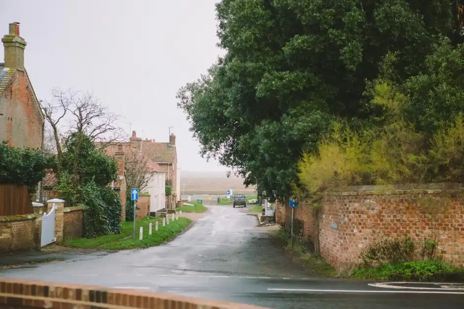 Village road in Burnham Overy Staithe, Norfolk, leading towards the coastal marshes, with traditional brick and flint cottages, mature trees and boundary walls.