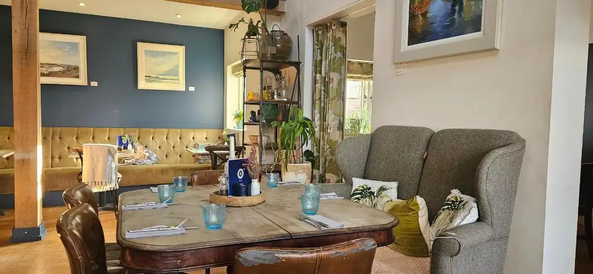 Restaurant interior at The Hero in Burnham Overy Staithe, Norfolk, featuring a wooden dining table, upholstered armchairs, leather seating and framed coastal artwork.