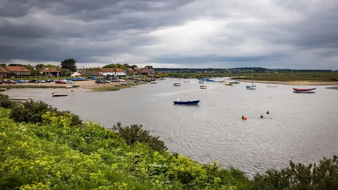 Burnham Overy Staithe Harbour on the Norfolk coast, with small sailing boats moored in the tidal creek, saltmarshes and village buildings beneath a cloudy sky.