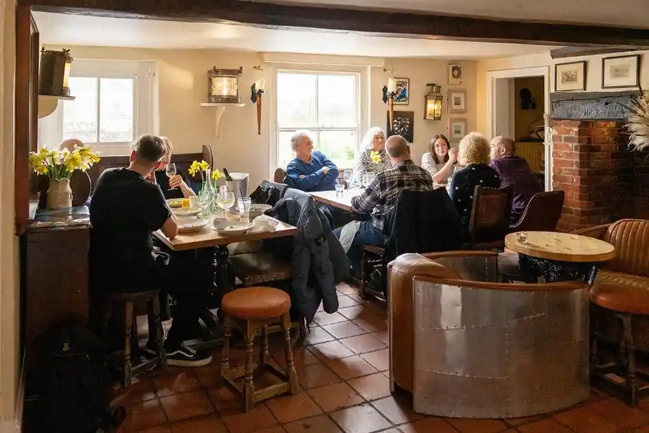 Traditional pub dining room at The Red Lion in Stiffkey, Norfolk, with guests seated around wooden tables, exposed ceiling beams, terracotta floor tiles and a brick fireplace.