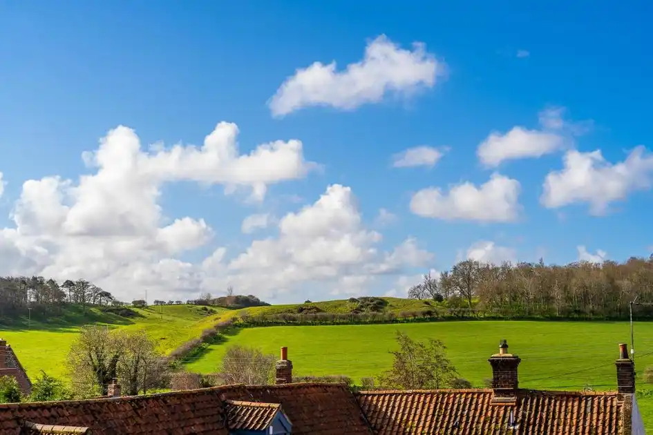 View across the rooftops of Stiffkey village in Norfolk towards rolling green fields, a countryside footpath and woodland beneath a blue sky with scattered clouds.