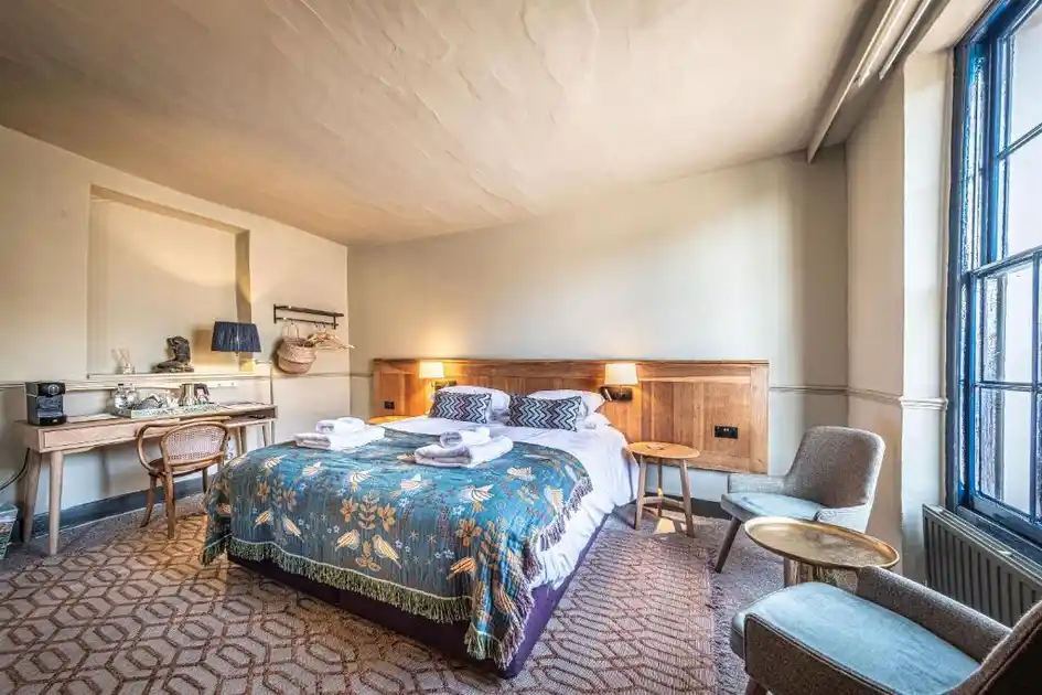 Guest bedroom at The Feathers in Holt, Norfolk, featuring a double bed with a patterned blue bedspread, wooden headboard, writing desk, upholstered chairs and a large sash window.