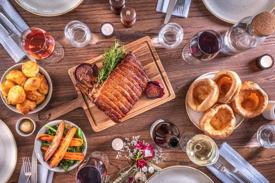 Roast dinner served at The Feathers in Holt, Norfolk, featuring sliced roast meat, Yorkshire puddings, roast potatoes, vegetables and glasses of wine on a wooden dining table.