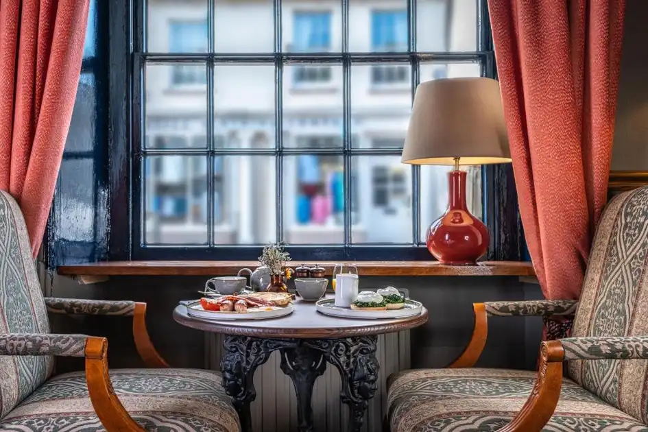 Breakfast served at a window-side table at The Feathers in Holt, Norfolk, featuring two upholstered armchairs, traditional leaded windows, red curtains and a table lamp.