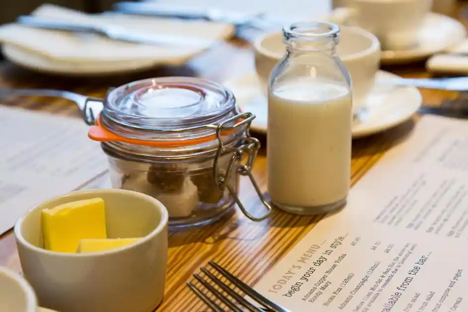 Breakfast table at The White Horse in Blakeney, Norfolk, featuring butter, a glass milk bottle, a jar of preserves, crockery and breakfast menus on a wooden table.