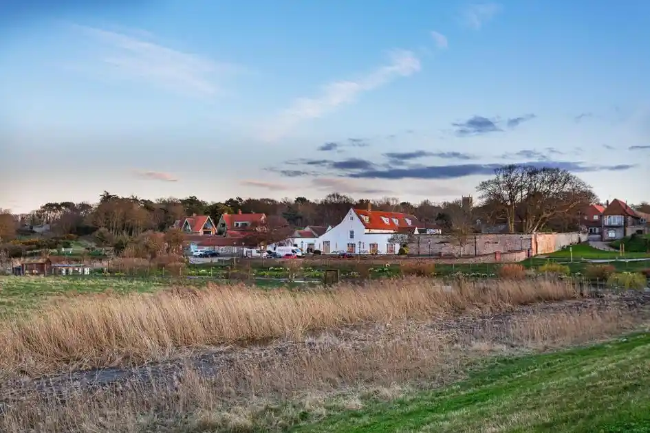 View across coastal saltmarshes towards The Manor Coastal Hotel & Inn in Blakeney, Norfolk, showing the white-painted hotel building, flint walls and surrounding village houses beneath an evening sky.