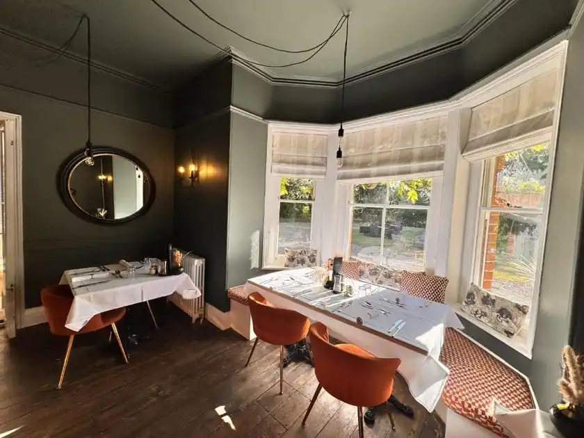 Restaurant dining room at Blakeney House in Blakeney, Norfolk, featuring tables set for dining, orange upholstered chairs, dark painted walls and large bay windows overlooking the garden.