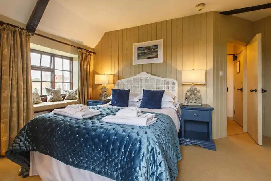 Guest bedroom at Cley Windmill in Cley-next-the-Sea, Norfolk, featuring a double bed with a blue quilted bedspread, wicker headboard, bedside lamps, exposed ceiling beams and a window seat.