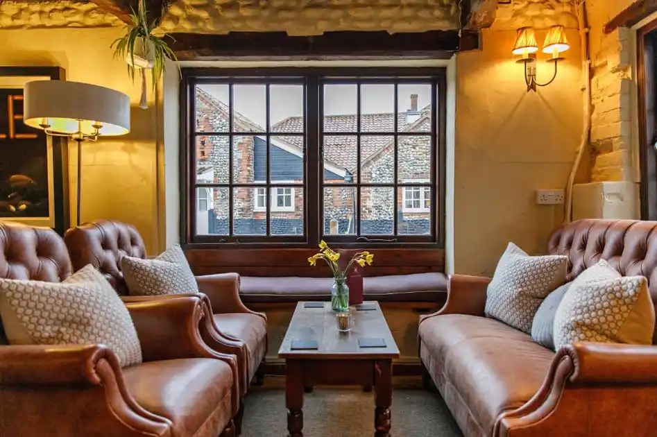 Guest lounge at Cley Windmill in Cley-next-the-Sea, Norfolk, featuring brown leather sofas and armchairs, exposed wooden beams, a coffee table and traditional windows overlooking flint-built village cottages.