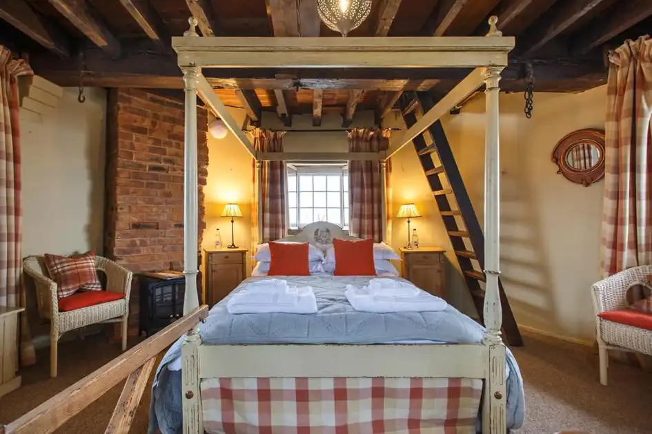 Characterful guest bedroom at Cley Windmill in Cley-next-the-Sea, Norfolk, featuring a white four-poster bed, exposed wooden ceiling beams, brickwork, tartan furnishings and a wooden staircase.