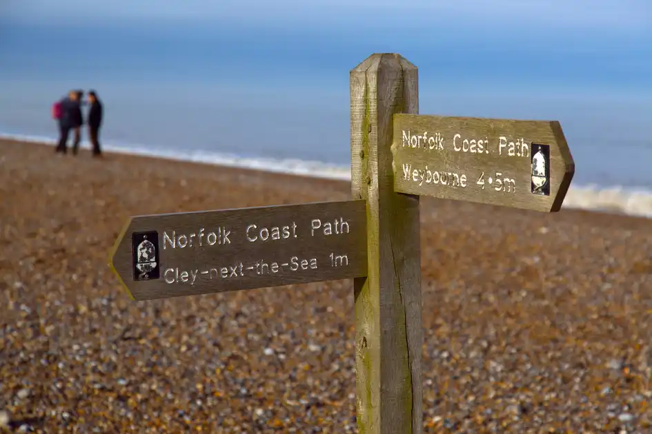Wooden Norfolk Coast Path signpost on the shingle beach at Cley-next-the-Sea, Norfolk, pointing towards Weybourne and Cley-next-the-Sea, with two walkers and the North Sea in the background.