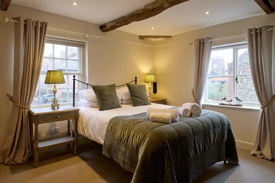 Guest bedroom at The Harnser in Cley-next-the-Sea, Norfolk, featuring a double bed with green bedspread, exposed wooden ceiling beams, bedside lamps, traditional sash windows and neatly arranged towels.