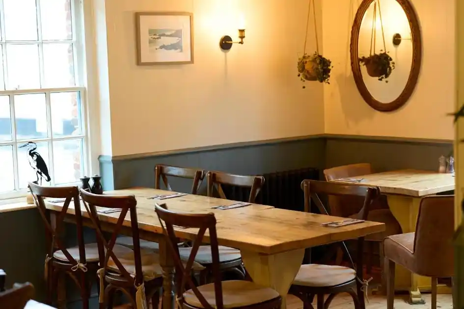 Bistro dining room at The Harnser in Cley-next-the-Sea, Norfolk, featuring wooden tables and chairs, traditional sash windows, grey wall panelling, hanging plants and a circular wall mirror.
