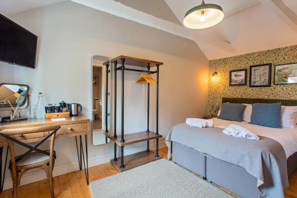 Guest bedroom at Three Swallows in Cley-next-the-Sea, Norfolk, featuring a double bed with grey bedspread, floral wallpaper, exposed wooden flooring, open clothes rail and tea and coffee-making facilities.