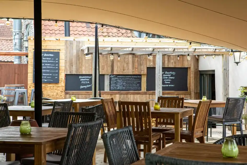 Covered outdoor dining area at Three Swallows in Cley-next-the-Sea, Norfolk, featuring wooden tables and chairs beneath a canopy, festoon lighting and a timber-clad pizza kitchen with chalkboard menus.