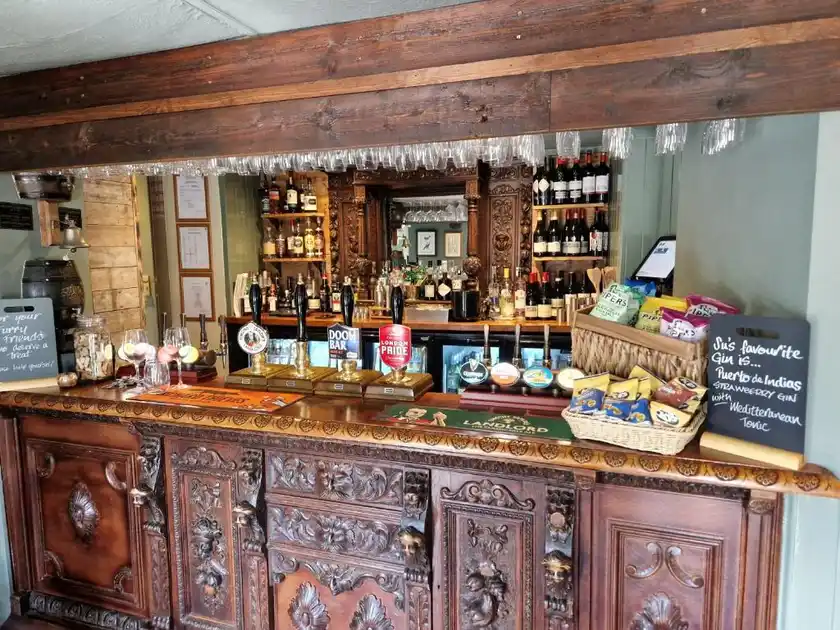 Traditional pub bar at Three Swallows in Cley-next-the-Sea, Norfolk, featuring an ornately carved wooden counter, beer pumps, shelves of wines and spirits, and exposed timber beams.