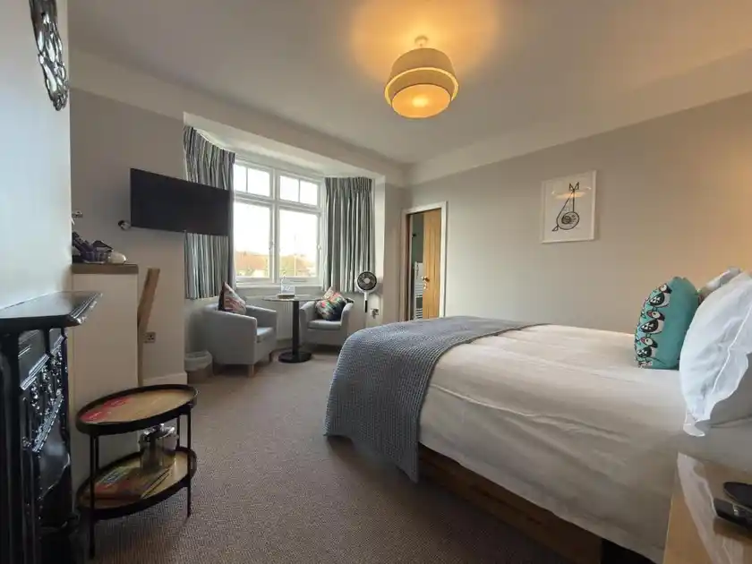 Guest bedroom at The Ship Inn in Weybourne, Norfolk, featuring a double bed with white bedding and blue throw, bay window with striped curtains, two upholstered armchairs, television and decorative fireplace.