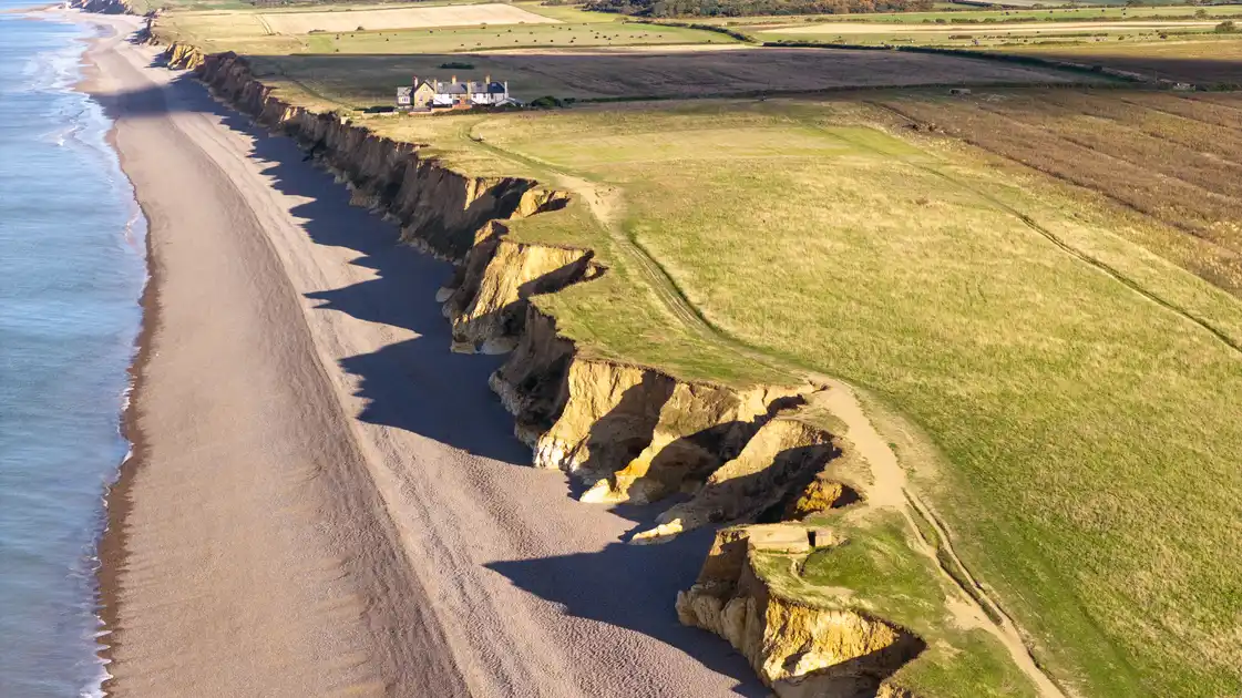Aerial view of Weybourne Beach on the North Norfolk coast, showing a broad shingle shoreline, eroding sandy cliffs, coastal footpaths and open farmland overlooking the North Sea.
