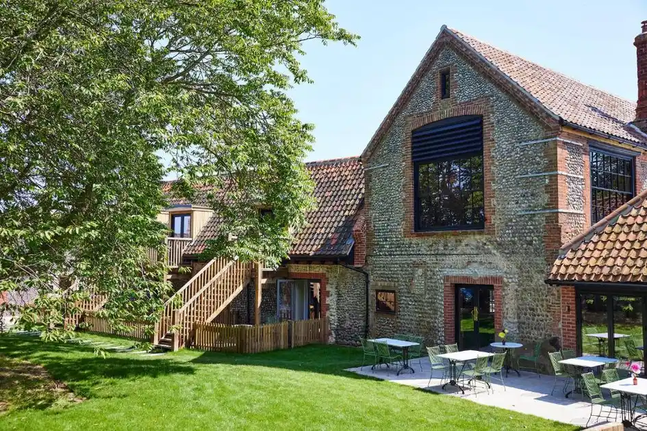 Exterior of The Maltings hotel in Weybourne, Norfolk, showing a restored flint and brick building with red pantile roofs, outdoor terrace seating, a green lawn and mature trees.