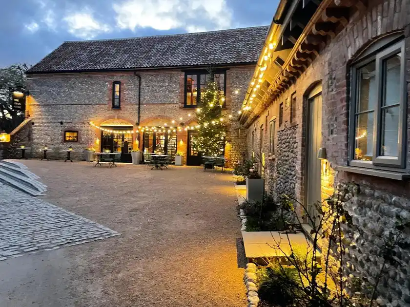 Evening view of the courtyard at The Maltings hotel in Weybourne, Norfolk, showing restored flint and brick buildings, illuminated outdoor seating, festoon lights and a decorated Christmas tree.