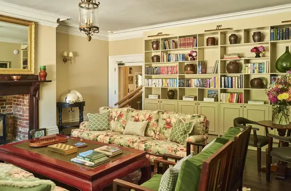 Library lounge at The Maltings hotel in Weybourne, Norfolk, featuring a floral sofa, green upholstered seating, bookshelves, brick fireplace, wooden coffee table and decorative lighting.