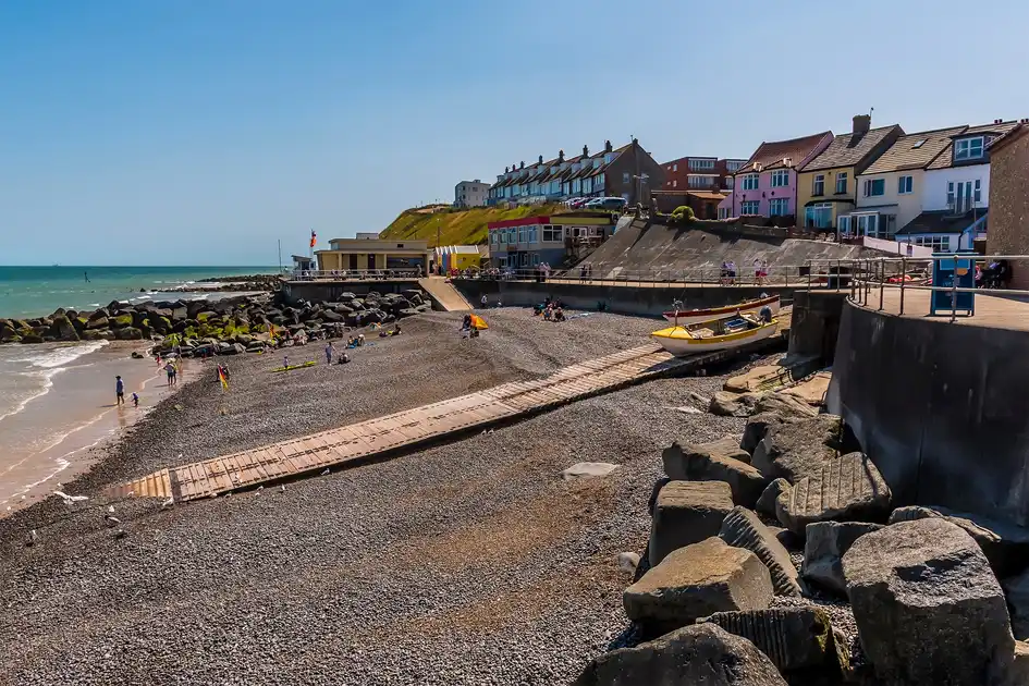 Sheringham Beach on the North Norfolk coast, showing the shingle shoreline, wooden boat ramp, fishing boats, sea defences and colourful seafront buildings overlooking the North Sea.