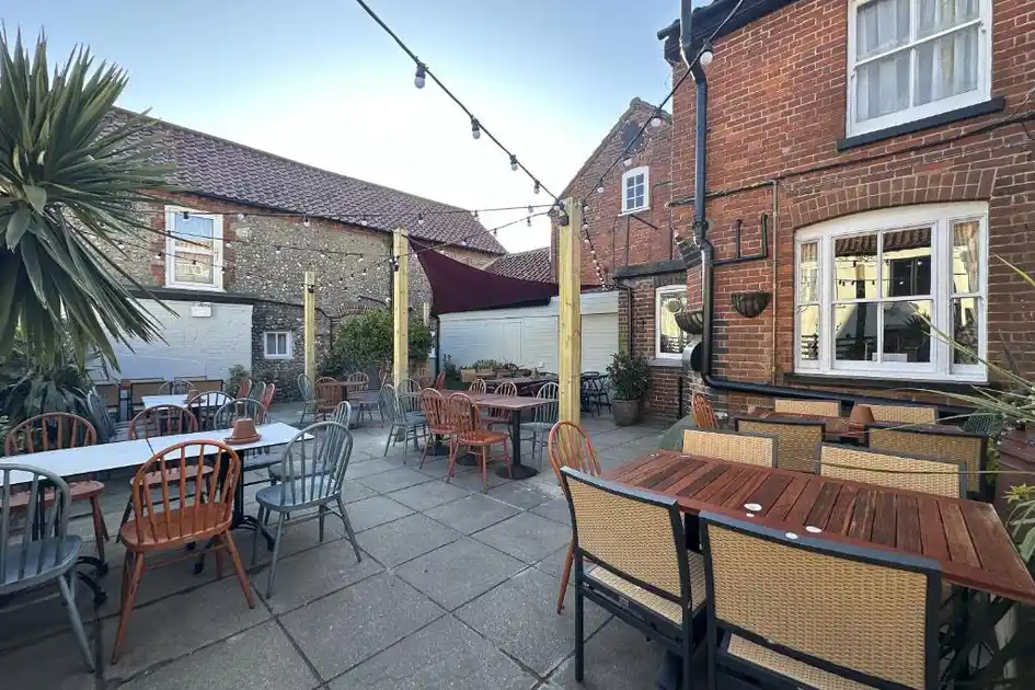 Outdoor beer garden at The Lobster in Sheringham, Norfolk, featuring wooden tables, mixed seating, festoon lighting and a paved courtyard enclosed by traditional brick and flint buildings.