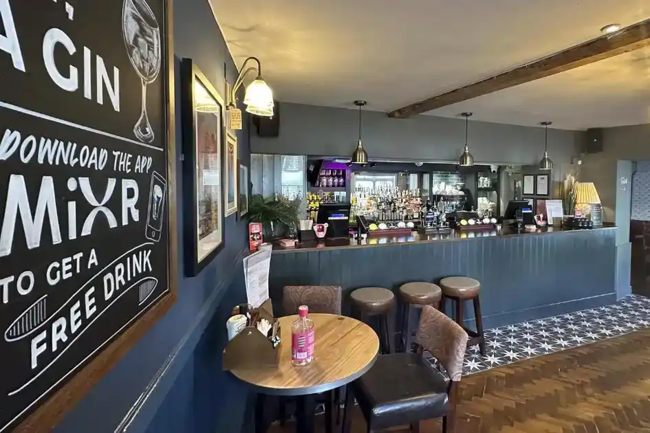 Interior of the bar at The Lobster in Sheringham, Norfolk, featuring a blue-painted bar counter, wooden stools, beer taps, pendant lighting, patterned floor tiles and tables for guests.