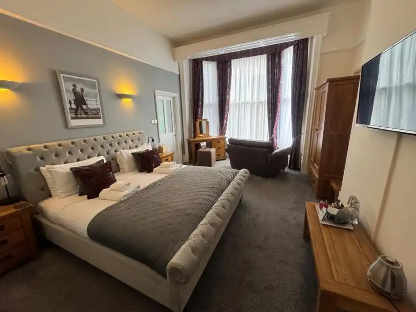 Spacious guest bedroom at The Red Lion Hotel in Cromer, Norfolk, featuring a large upholstered double bed, grey bedding, high ceilings, bay windows, seating area and wooden furnishings.