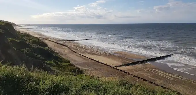 Overstrand Beach on the Norfolk coast, showing a sandy shoreline, timber groynes, grassy coastal cliffs and waves breaking along the North Sea coast.